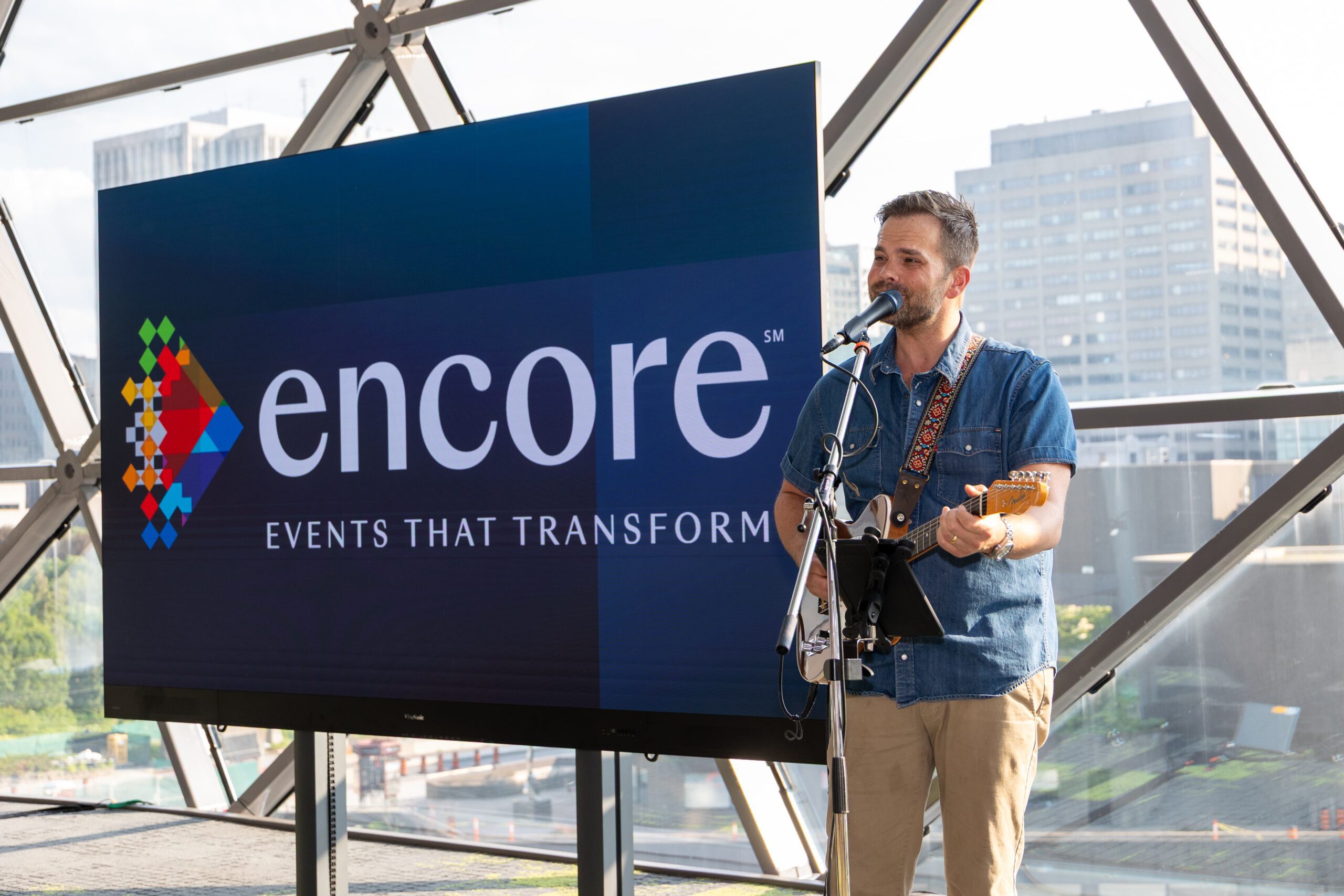 Man plays electric guitar and sings into a microphone beside a large "Encore Events That Transform" display screen.