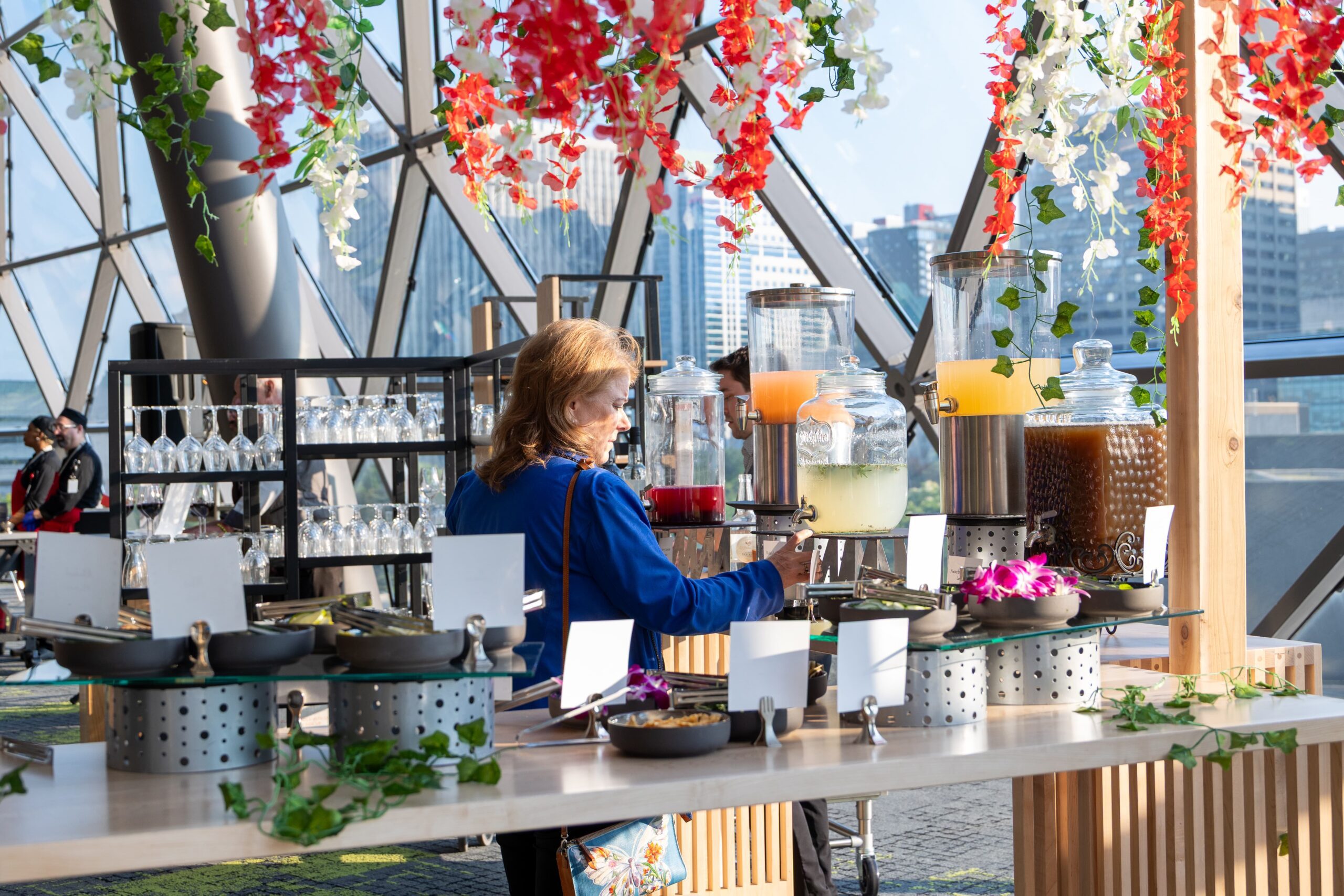 A woman serves herself a drink at a self-serve beverage station with dispensers and hanging flowers.