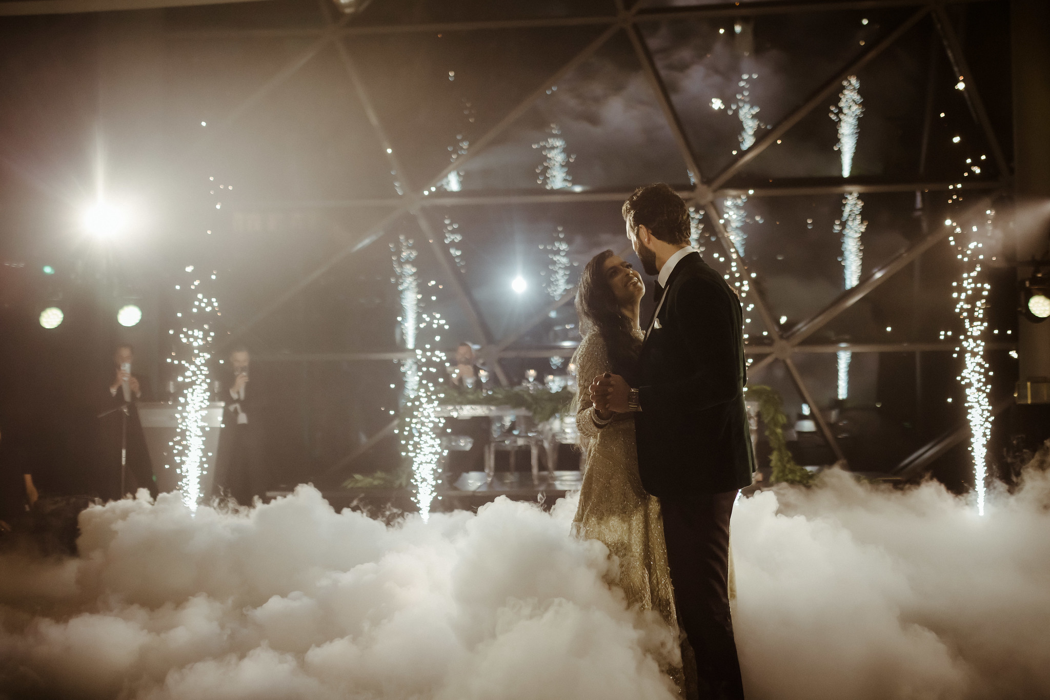 A couple dances among fog and indoor fireworks, with bright lights and musicians visible in the background.