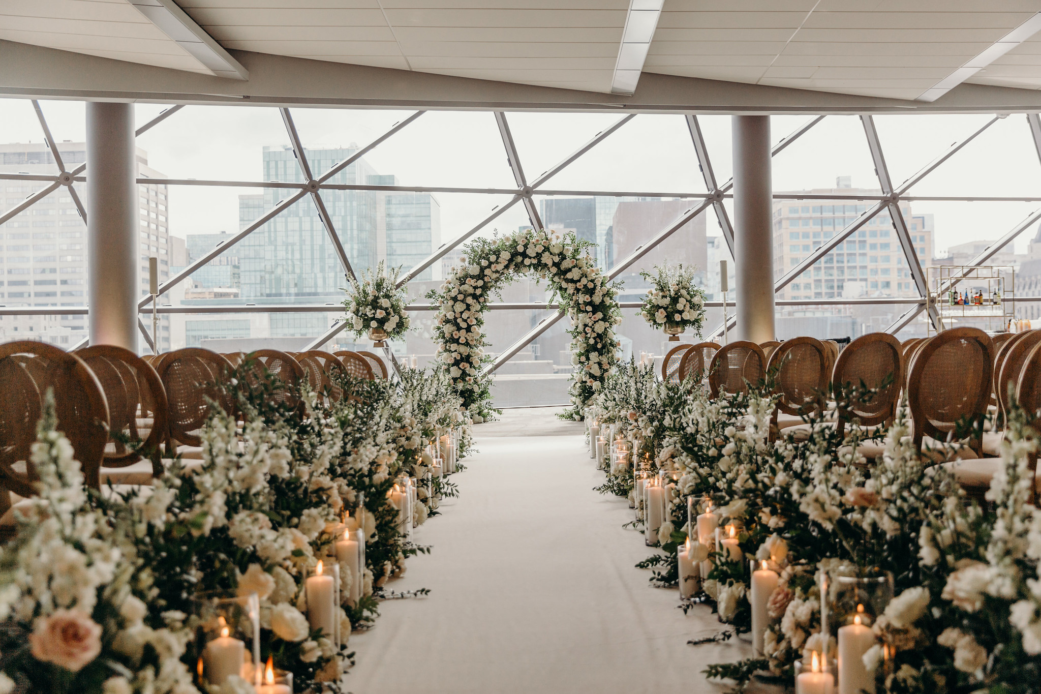 Indoor wedding aisle with floral arrangements, candles, and a white flower arch, set against large city windows.