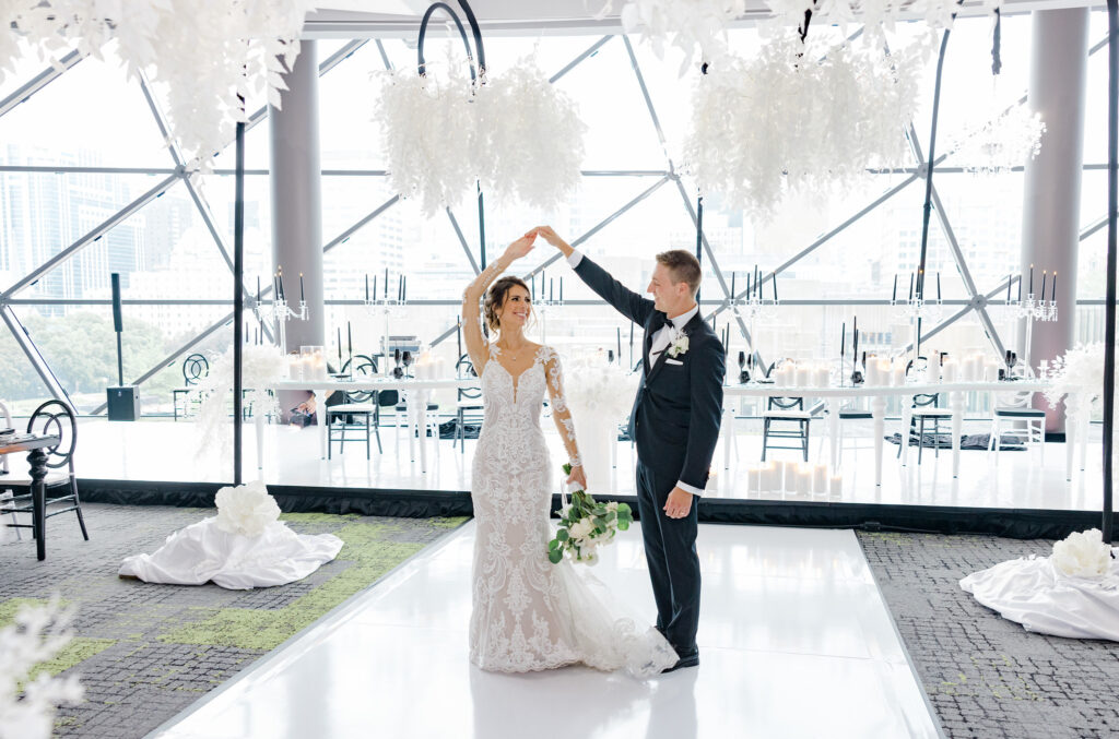 Bride and groom dance together in a modern, white-decorated venue with large windows and floral arrangements.