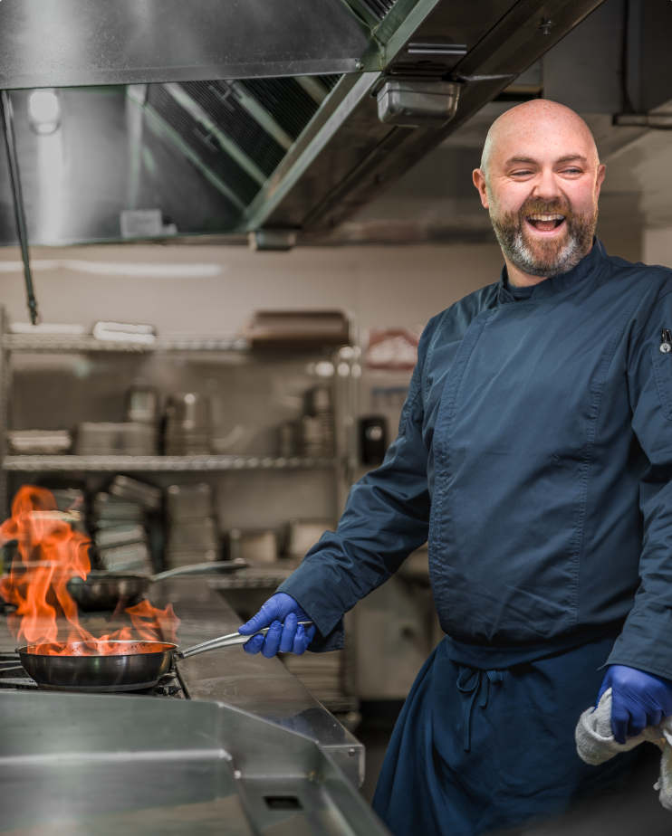 Chef in a navy uniform cooks with a flaming pan in a commercial kitchen, smiling and wearing blue gloves.