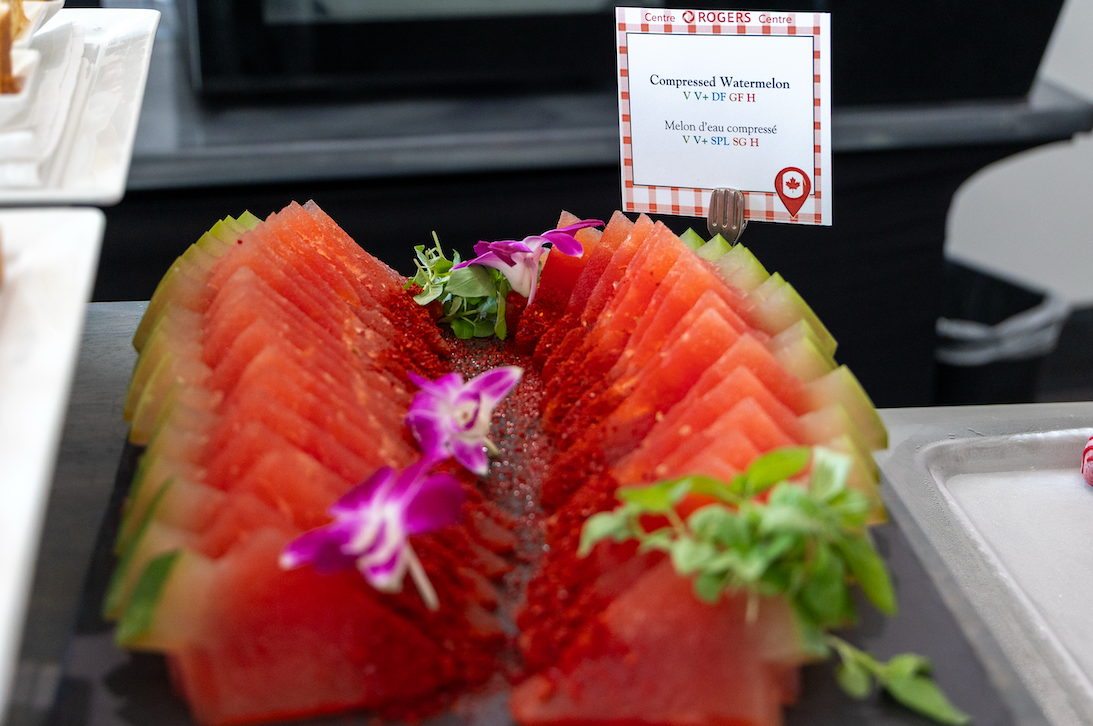 Sliced compressed watermelon garnished with edible flowers and herbs, labeled with a sign featuring dietary information.
