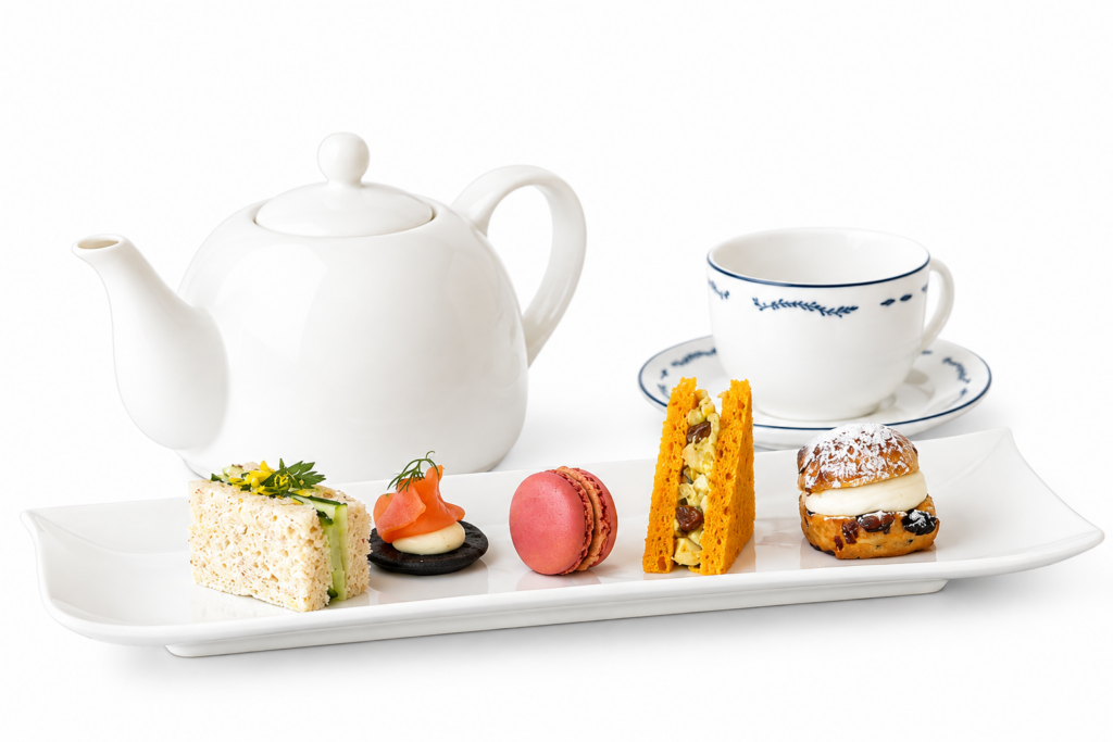 White teapot and cup with assorted pastries, sandwich, and macaron arranged on a rectangular white plate.