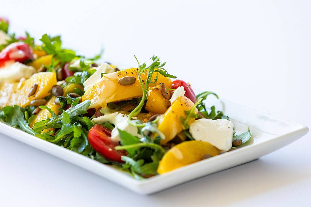 Salad with arugula, tomatoes, feta cheese, yellow peaches, and pumpkin seeds on a rectangular white plate.