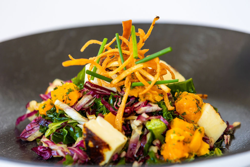 Mixed salad with greens, grilled cheese, cubed squash, shredded carrots, and chives on a black plate.