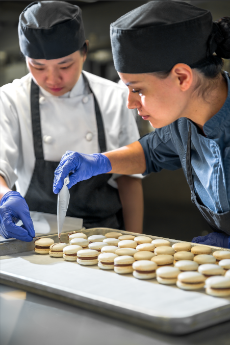 Two chefs in uniforms and gloves are piping filling onto rows of macarons on a baking tray in a kitchen.